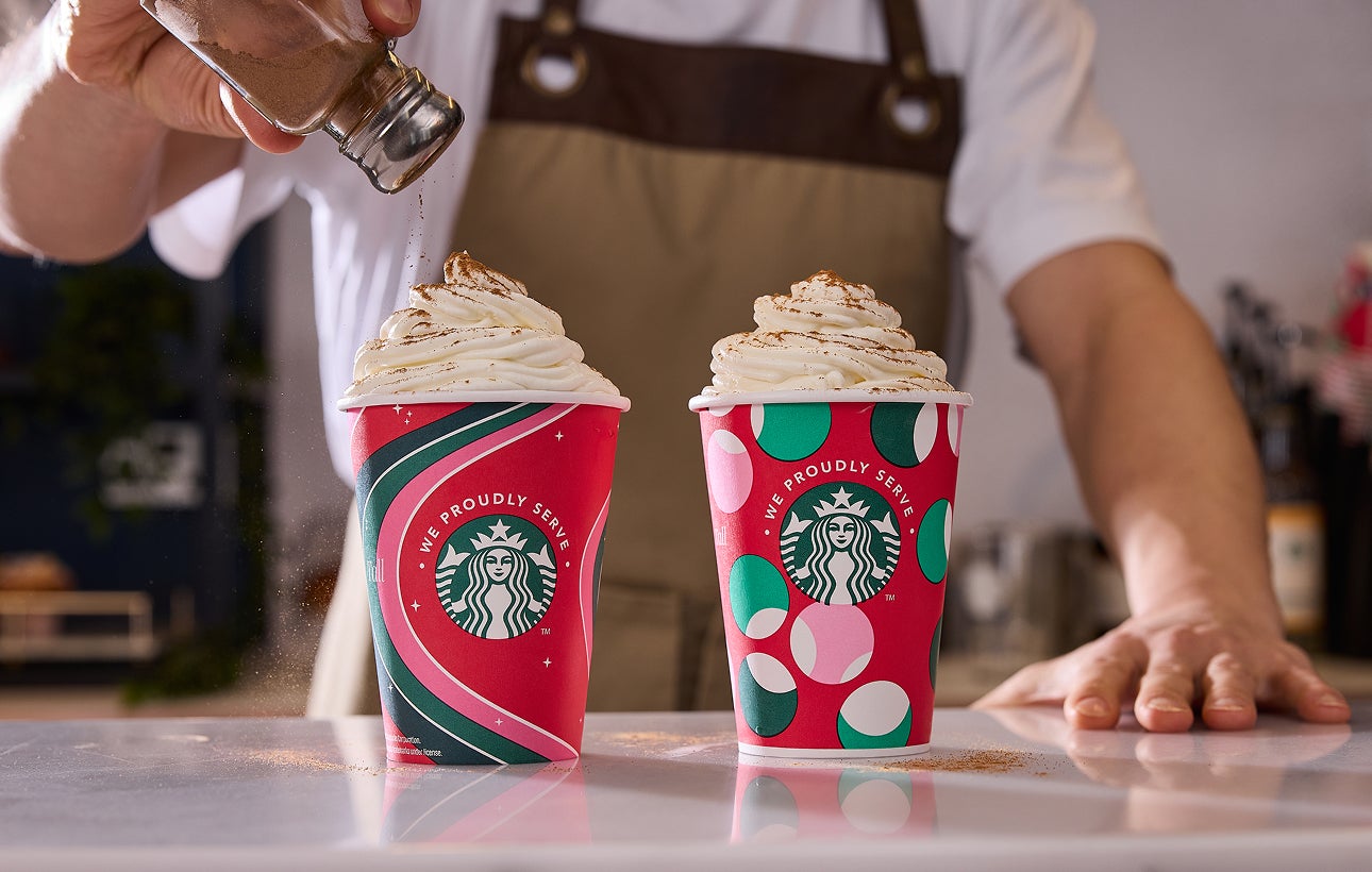 Două băuturi Frappuccino® Blended în căni festive We Proudly Serve Starbucks, cu scorțișoară presărată peste una dintre ele