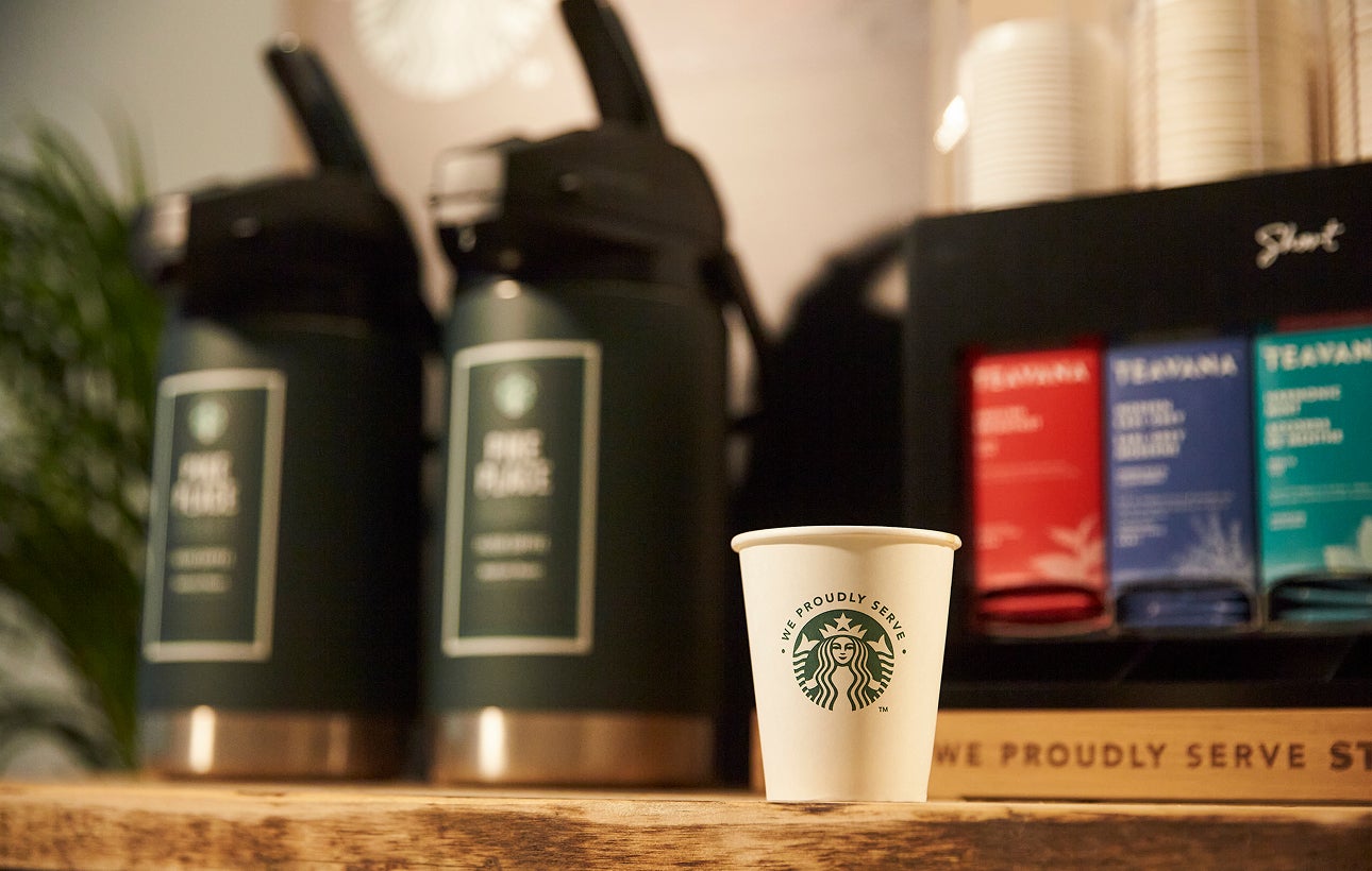 Un pahar de hârtie We Proudly Serve Starbucks se află pe un blat de lemn în fața unor dozatoare mari de cafea preparată