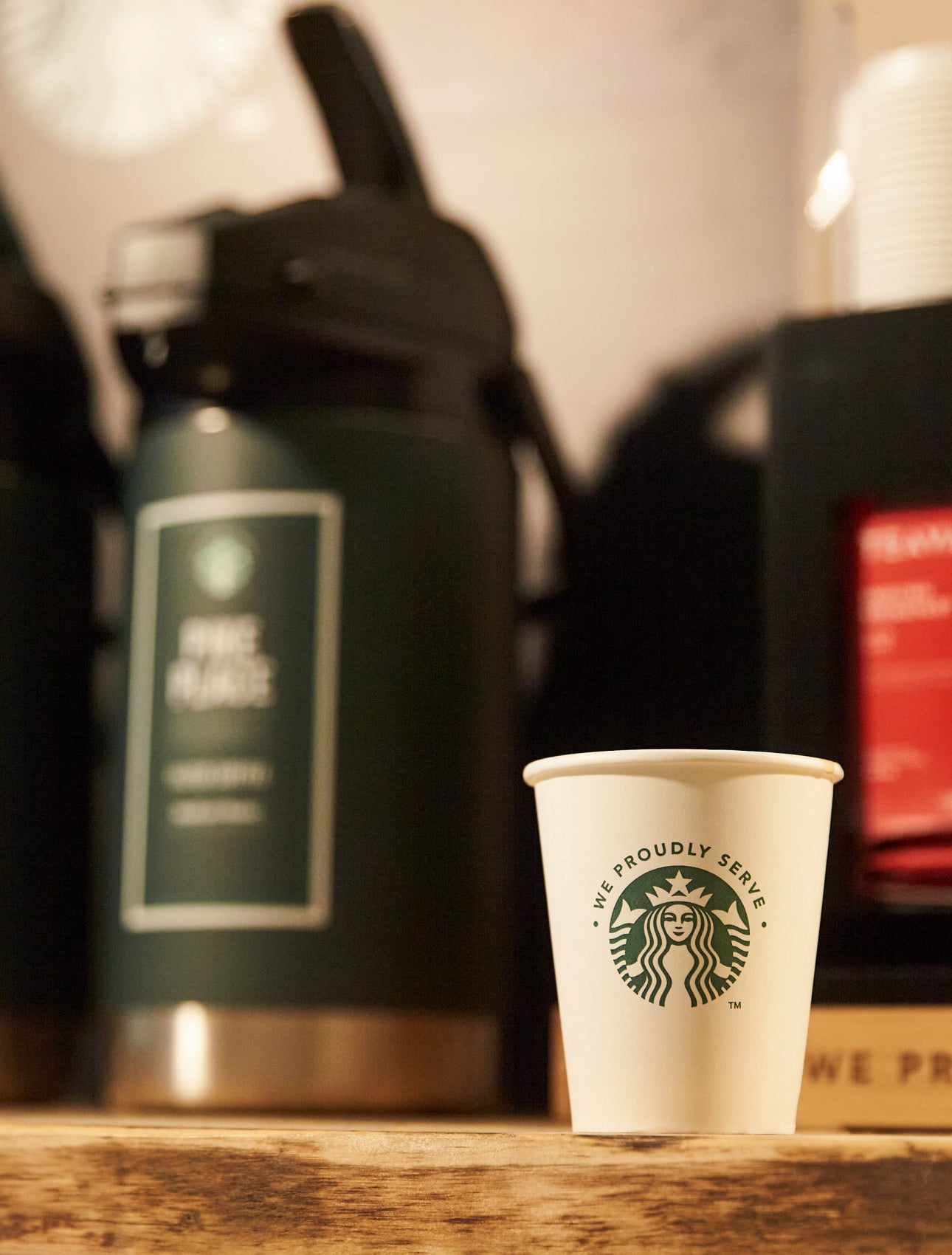 Un pahar de hârtie We Proudly Serve Starbucks se află pe un blat de lemn în fața unor dozatoare mari de cafea preparată