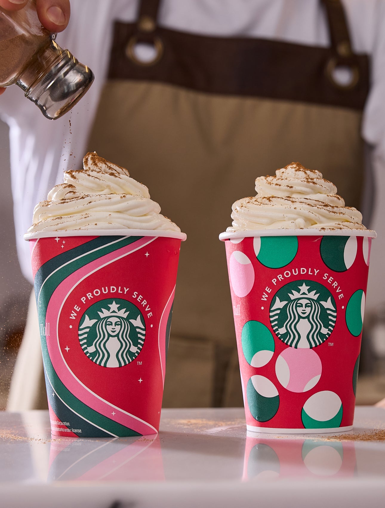 Două băuturi Frappuccino® Blended în căni festive We Proudly Serve Starbucks, cu scorțișoară presărată peste una dintre ele