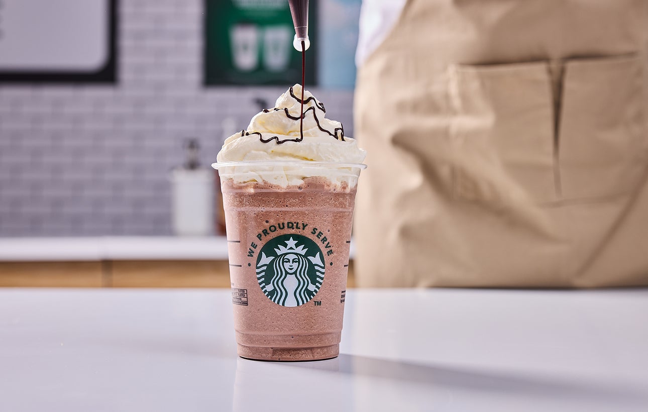 O băutură We Proudly Serve Starbucks, acoperită cu frișcă, iar un barista adaugă deasupra un strop de ciocolată.