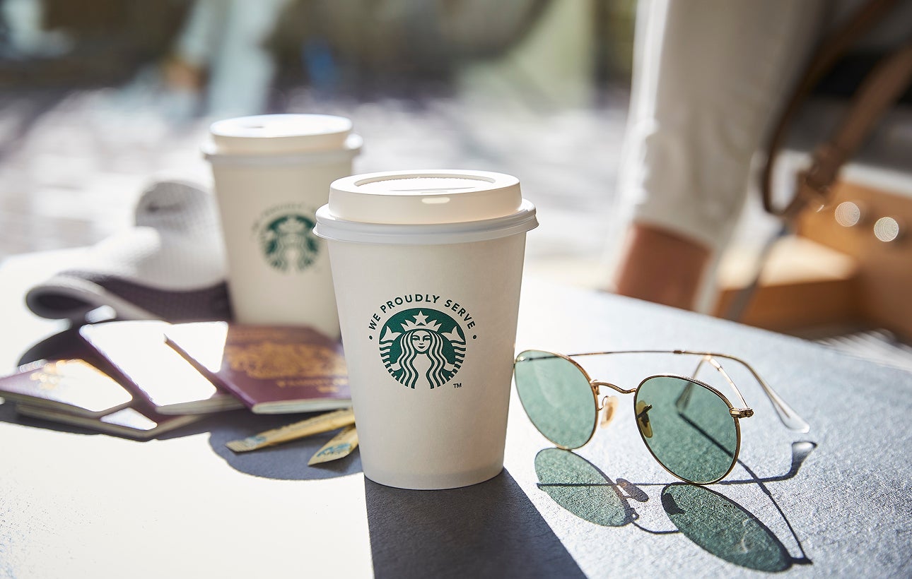 Două pahare cu capac We Proudly Serve Starbucks, așezate pe o masă în aer liber, lângă o pereche de ochelari de soare