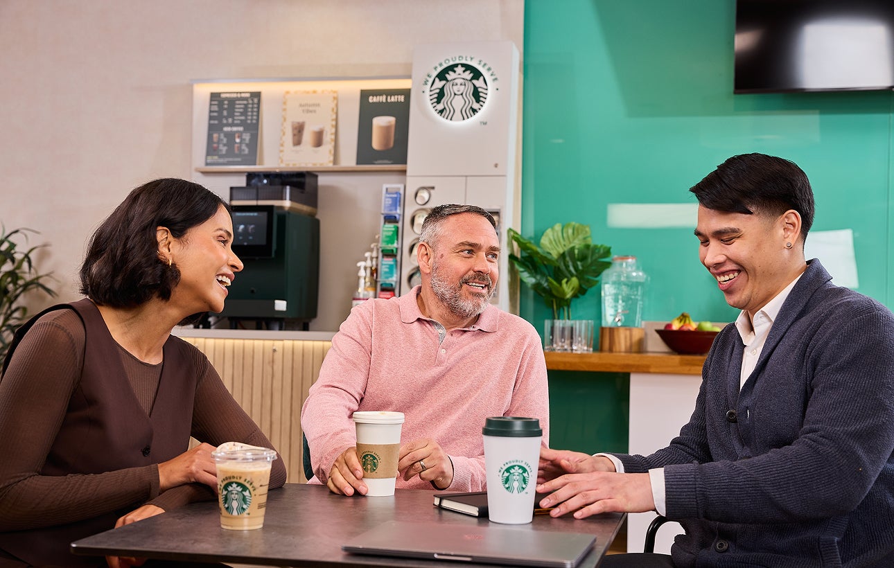 Colegi stând împreună cu ceștile „We Proudly Serve Starbucks” și un aparat de cafea cu autoservire în fundal