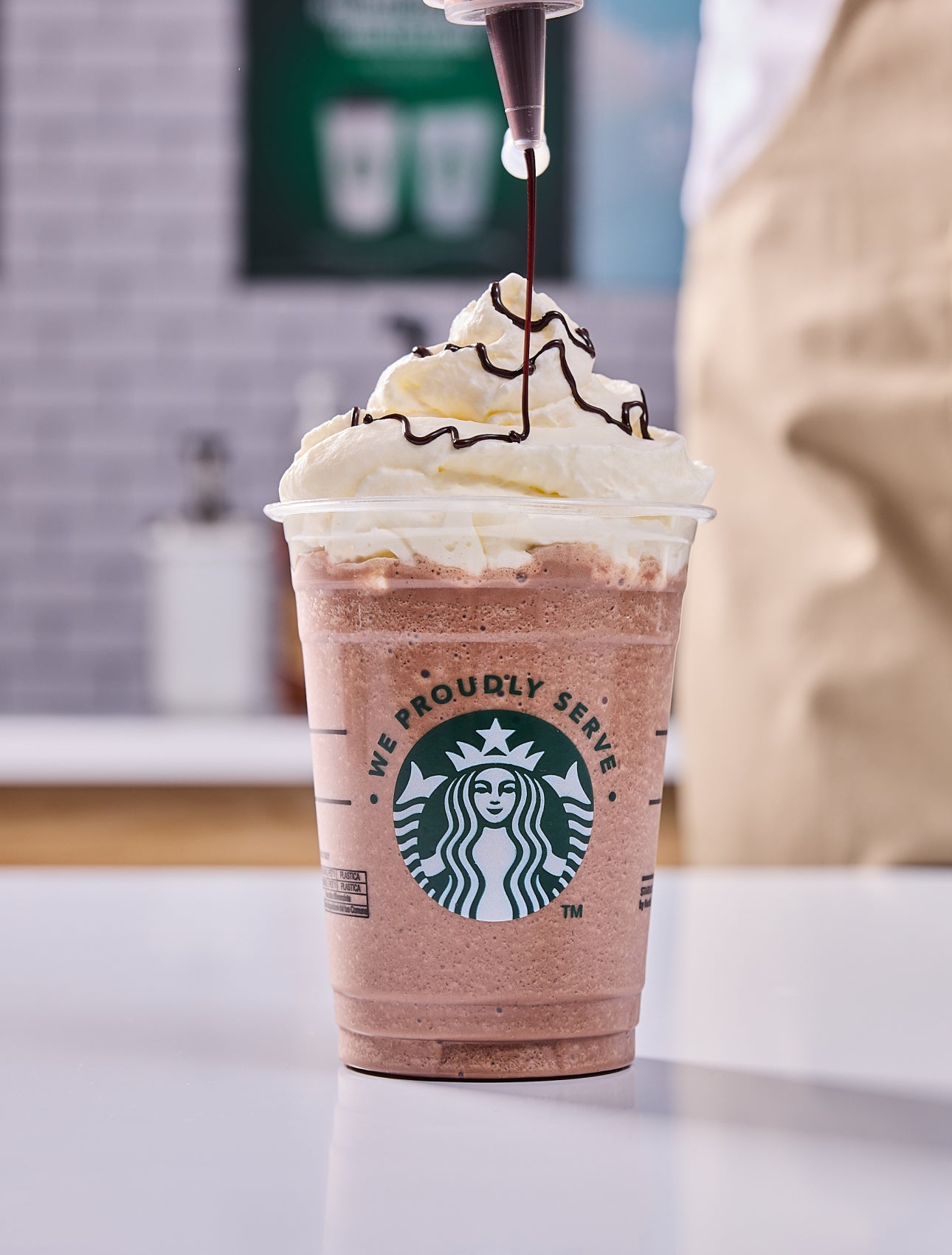 O băutură We Proudly Serve Starbucks, acoperită cu frișcă, iar un barista adaugă deasupra un strop de ciocolată.