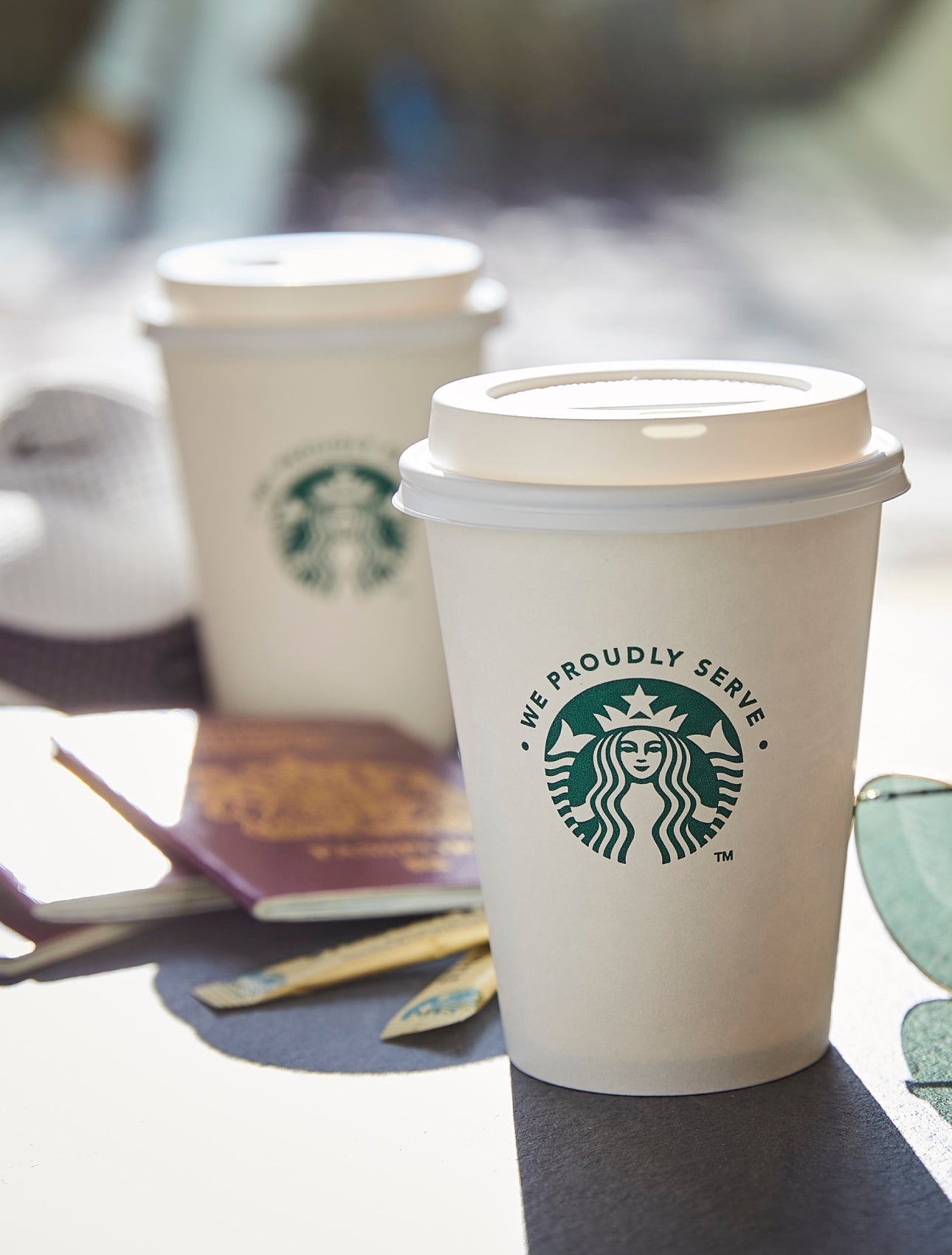 Două pahare cu capac We Proudly Serve Starbucks, așezate pe o masă în aer liber, lângă o pereche de ochelari de soare