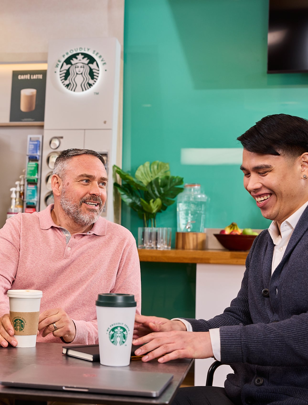 Colegi stând împreună cu ceștile „We Proudly Serve Starbucks” și un aparat de cafea cu autoservire în fundal