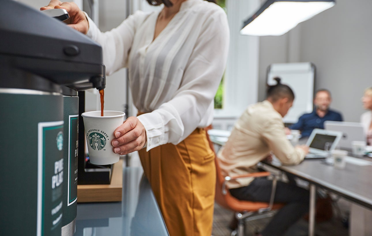O persoană umple un pahar de hârtie We Proudly Serve Starbucks cu cafea preparată la un dozator de cafea comercial