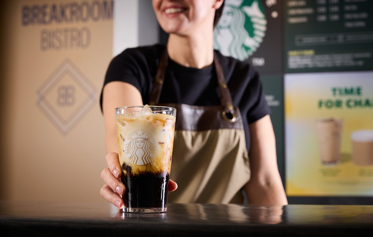 Un barista servește un pahar We Proudly Serve Starbucks umplut cu cafea cu gheață, prezentând straturi de cafea și cremă
