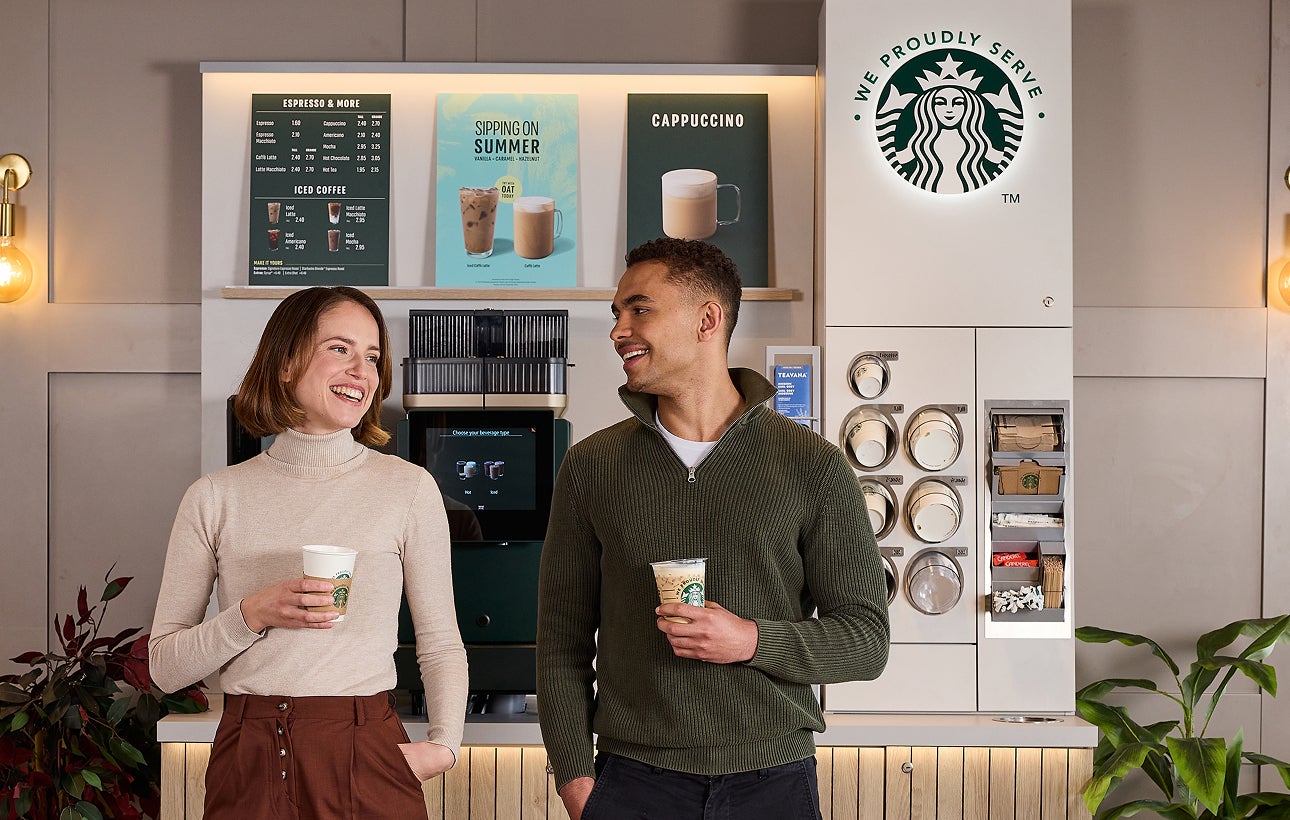 Două persoane care stau de vorbă în timp ce țin în mână cești de cafea cu mesajul „We Proudly Serve Starbucks” în fața unui a