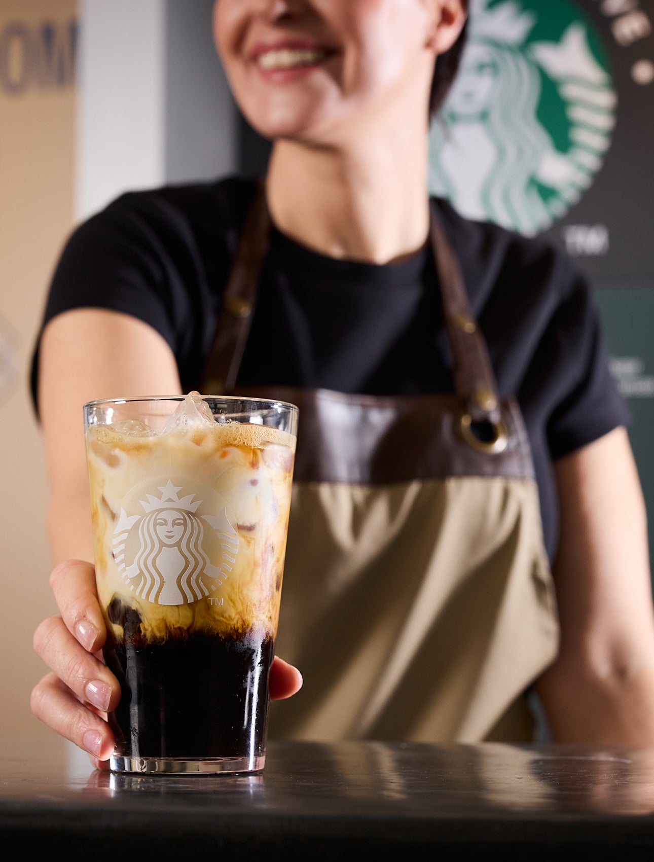 Un barista servește un pahar We Proudly Serve Starbucks umplut cu cafea cu gheață, prezentând straturi de cafea și cremă