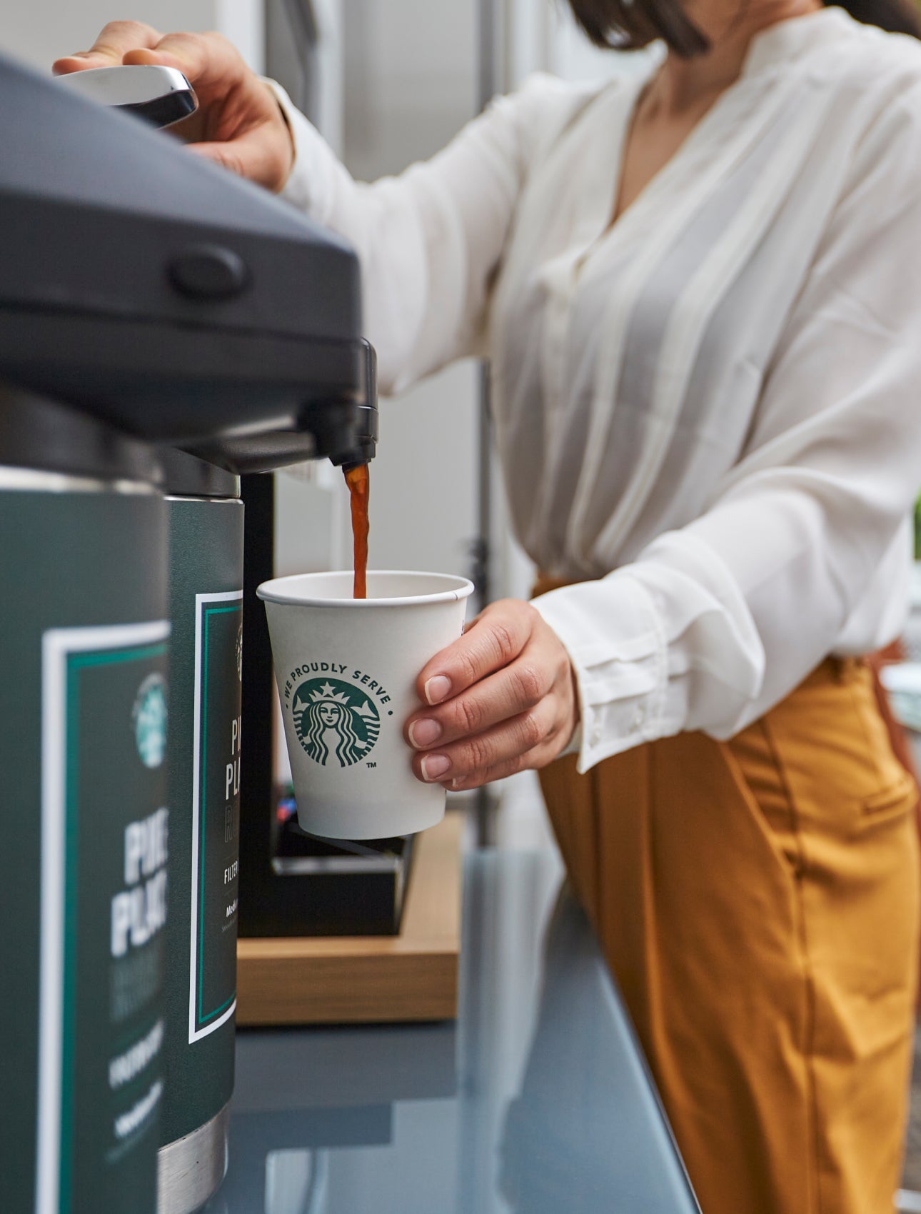 O persoană umple un pahar de hârtie We Proudly Serve Starbucks cu cafea preparată la un dozator de cafea comercial