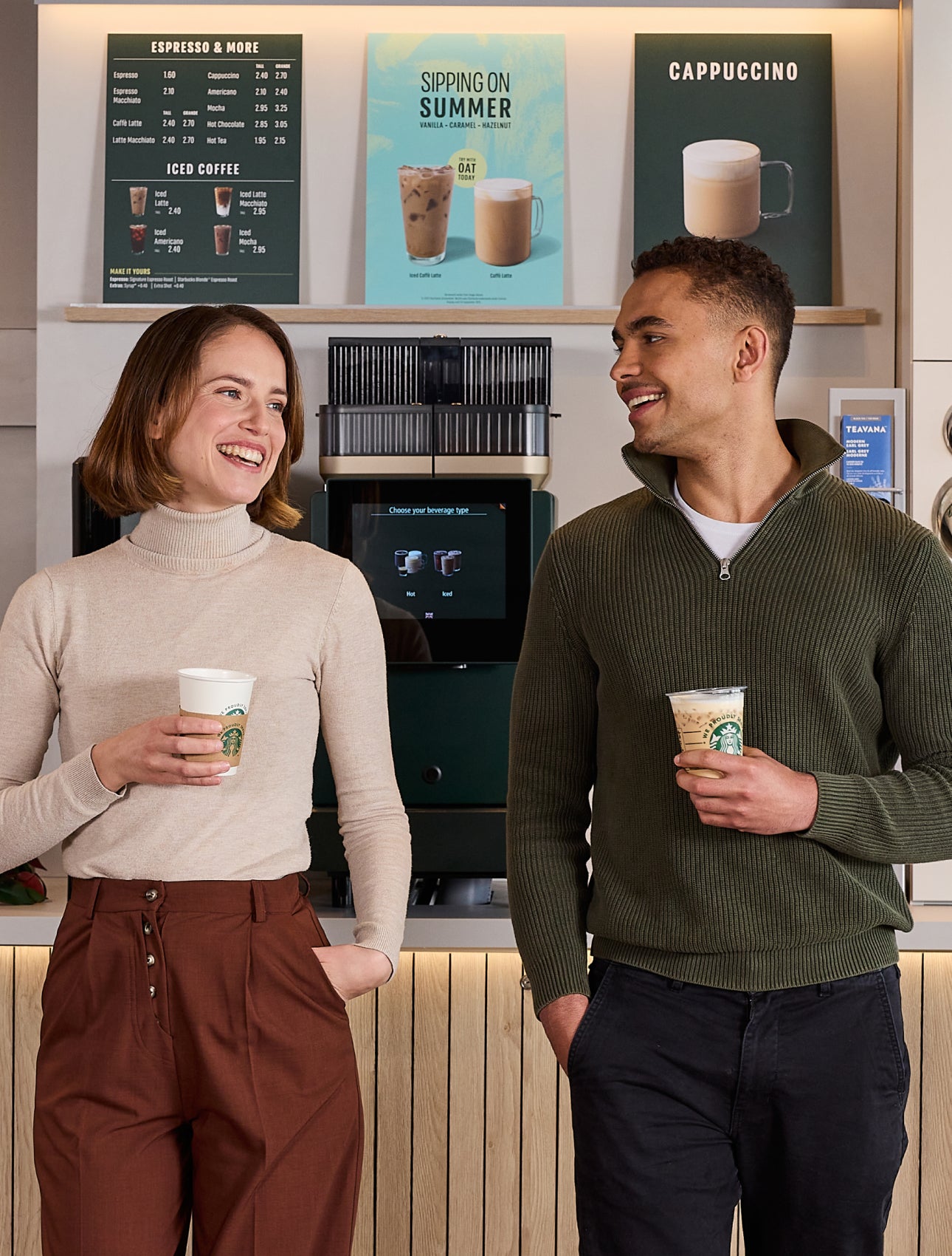 Două persoane care stau de vorbă în timp ce țin în mână cești de cafea cu mesajul „We Proudly Serve Starbucks” în fața unui a