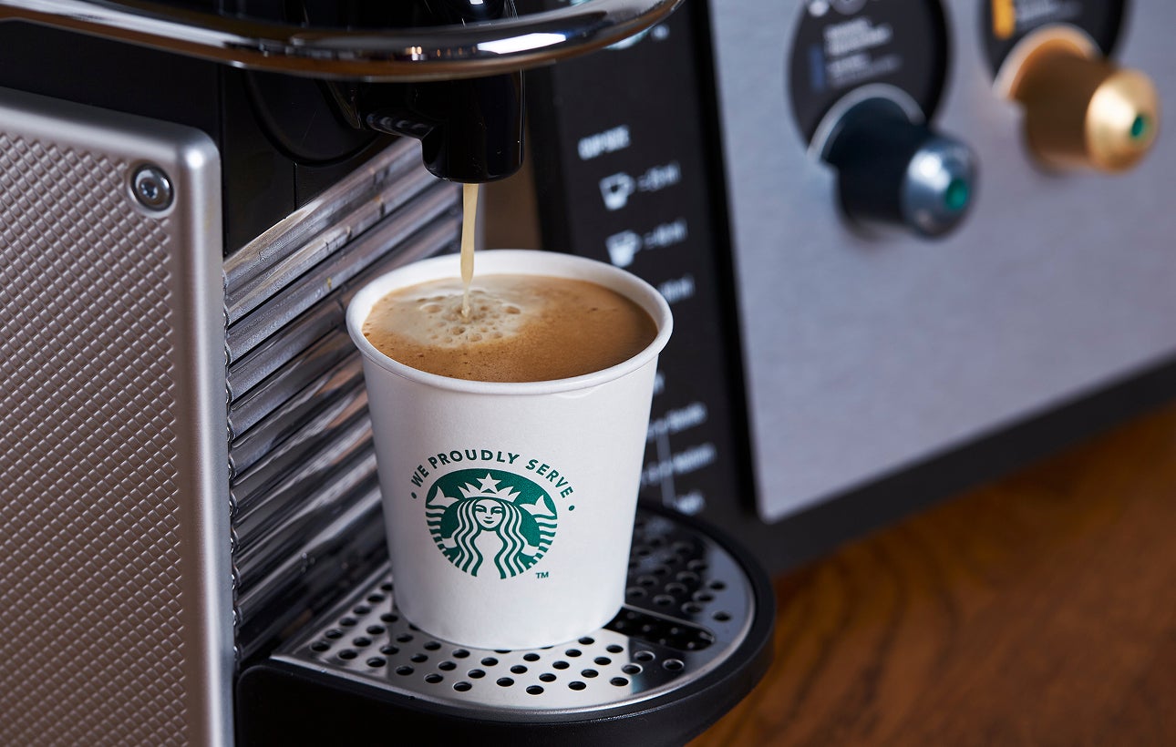 Un pahar de hârtie We Proudly Serve Starbucks plin cu cafea proaspăt preparată de la un espressor 