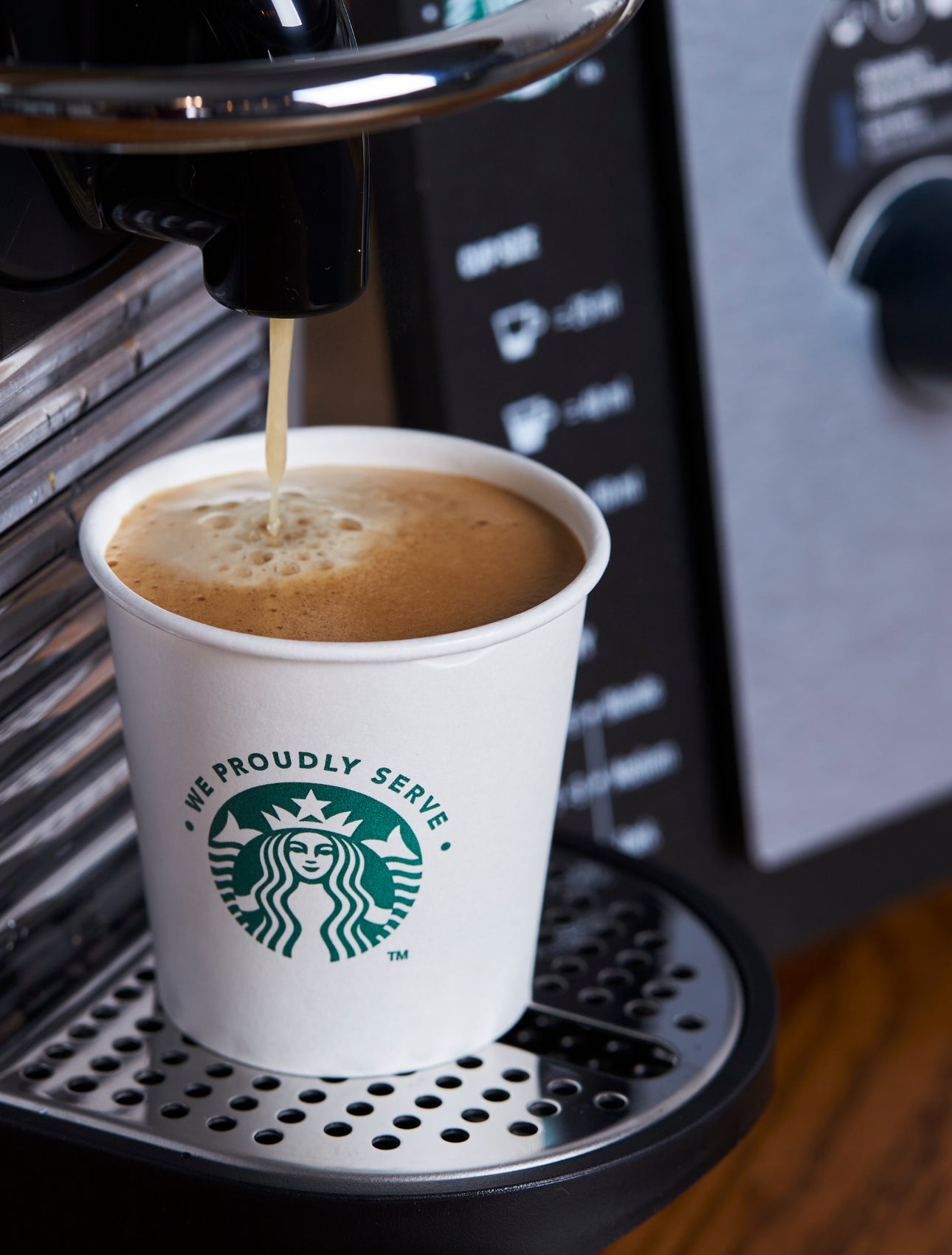 Un pahar de hârtie We Proudly Serve Starbucks plin cu cafea proaspăt preparată de la un espressor 
