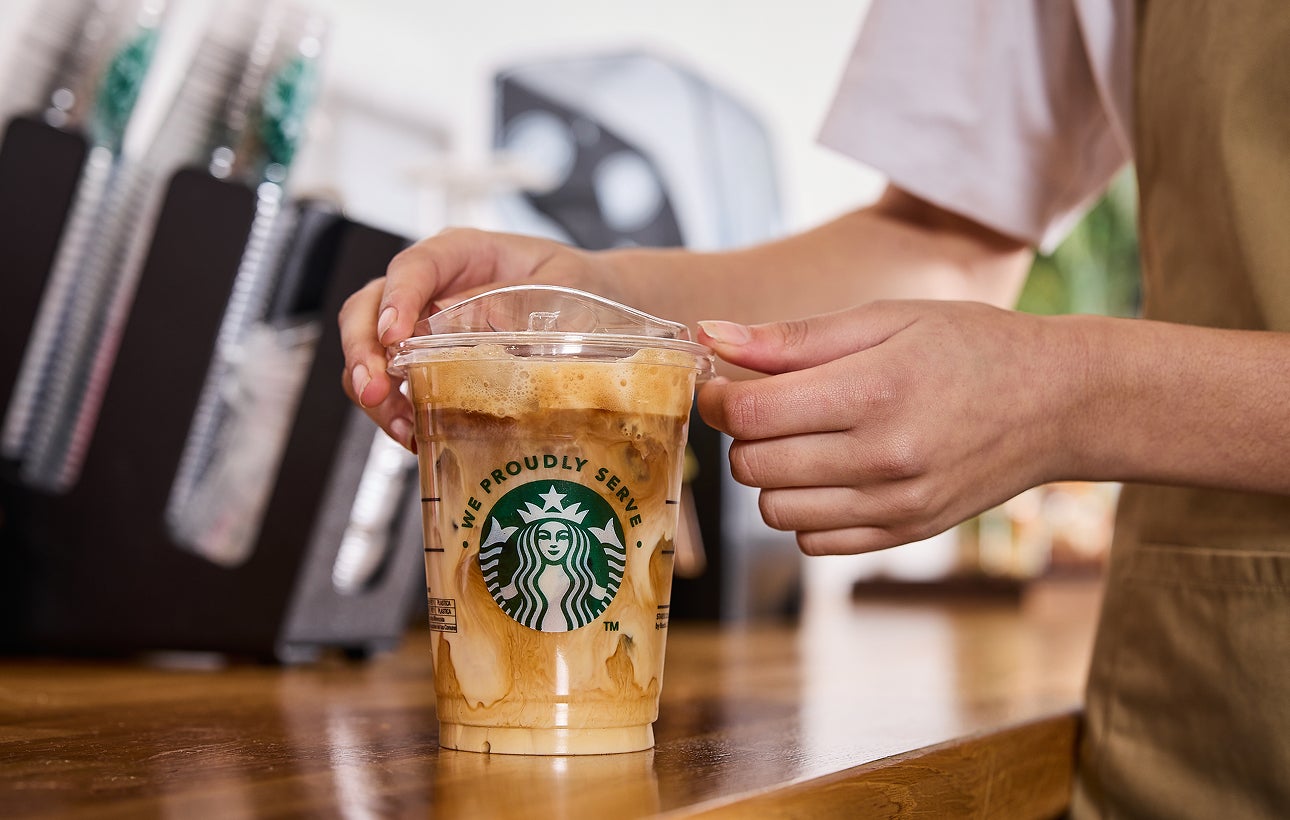 Un barista pune capacul pe o cană de cafea cu gheață We Proudly Serve Starbucks, cu spirale de cremă și cafea