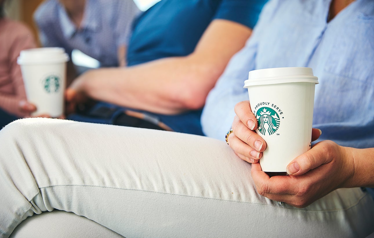 O persoană așezată, ținând cu ambele mâini un pahar de cafea We Proudly Serve Starbucks la pachet.