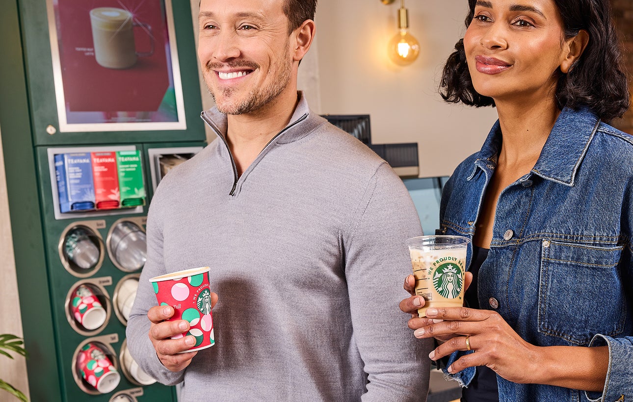 Un bărbat care ține o ceașcă festivă de cafea „We Proudly Serve Starbucks” și o femeie care ține o cafea cu gheață