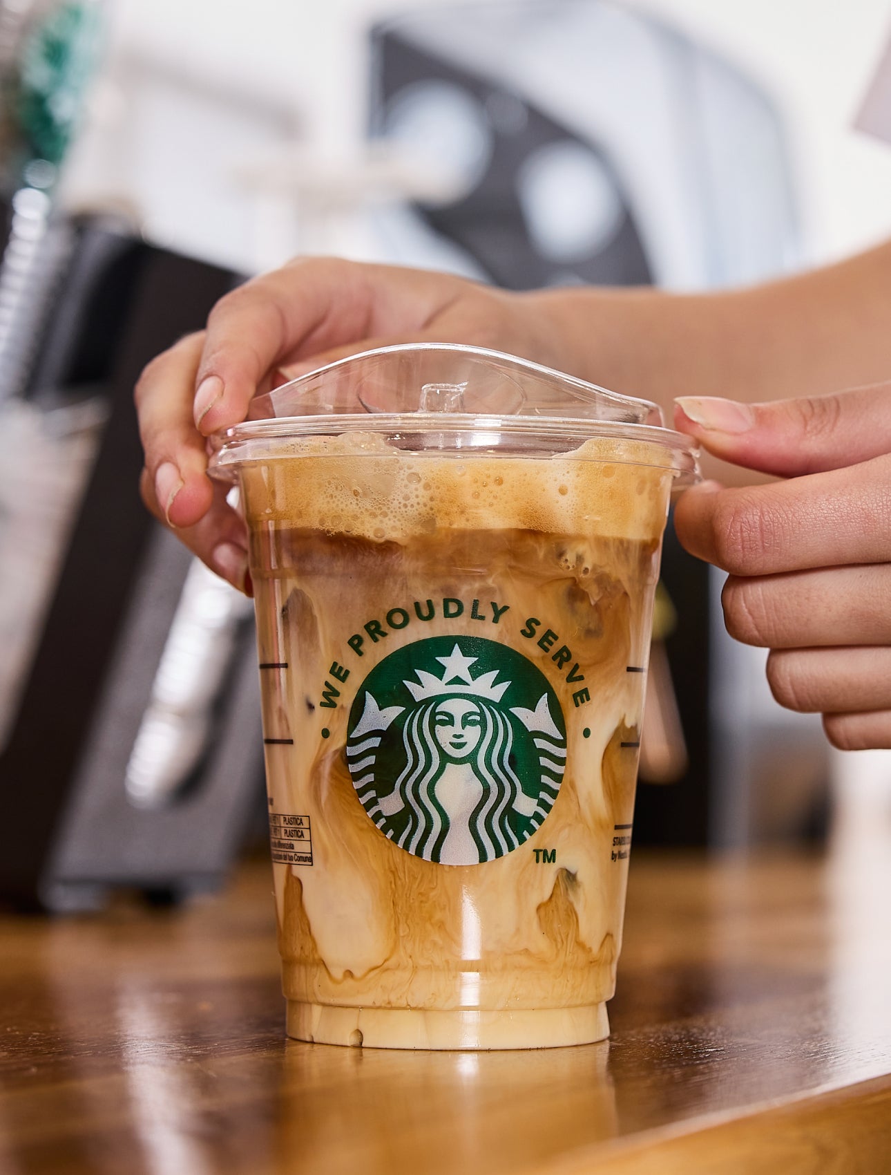 Un barista pune capacul pe o cană de cafea cu gheață We Proudly Serve Starbucks, cu spirale de cremă și cafea