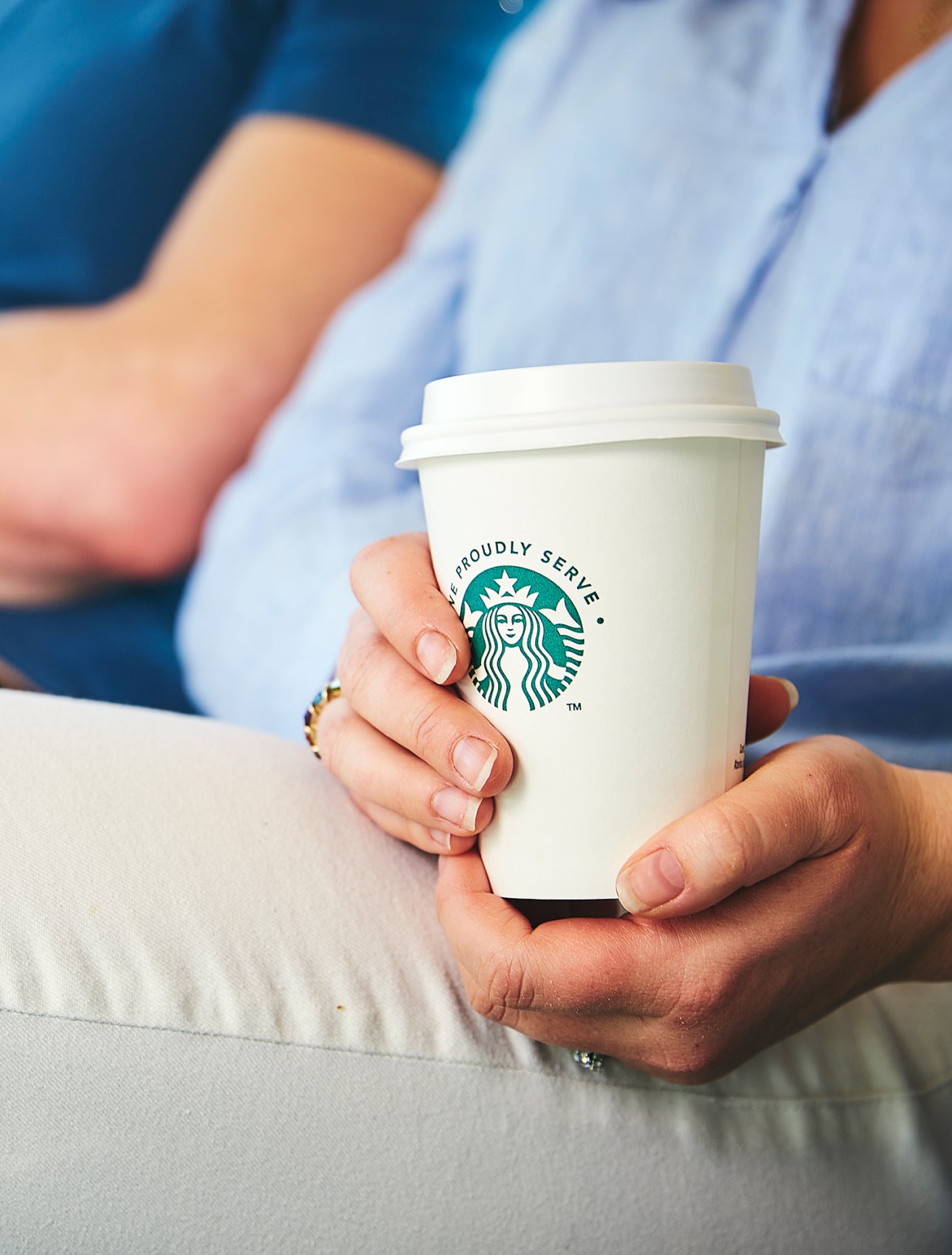 O persoană așezată, ținând cu ambele mâini un pahar de cafea We Proudly Serve Starbucks la pachet.