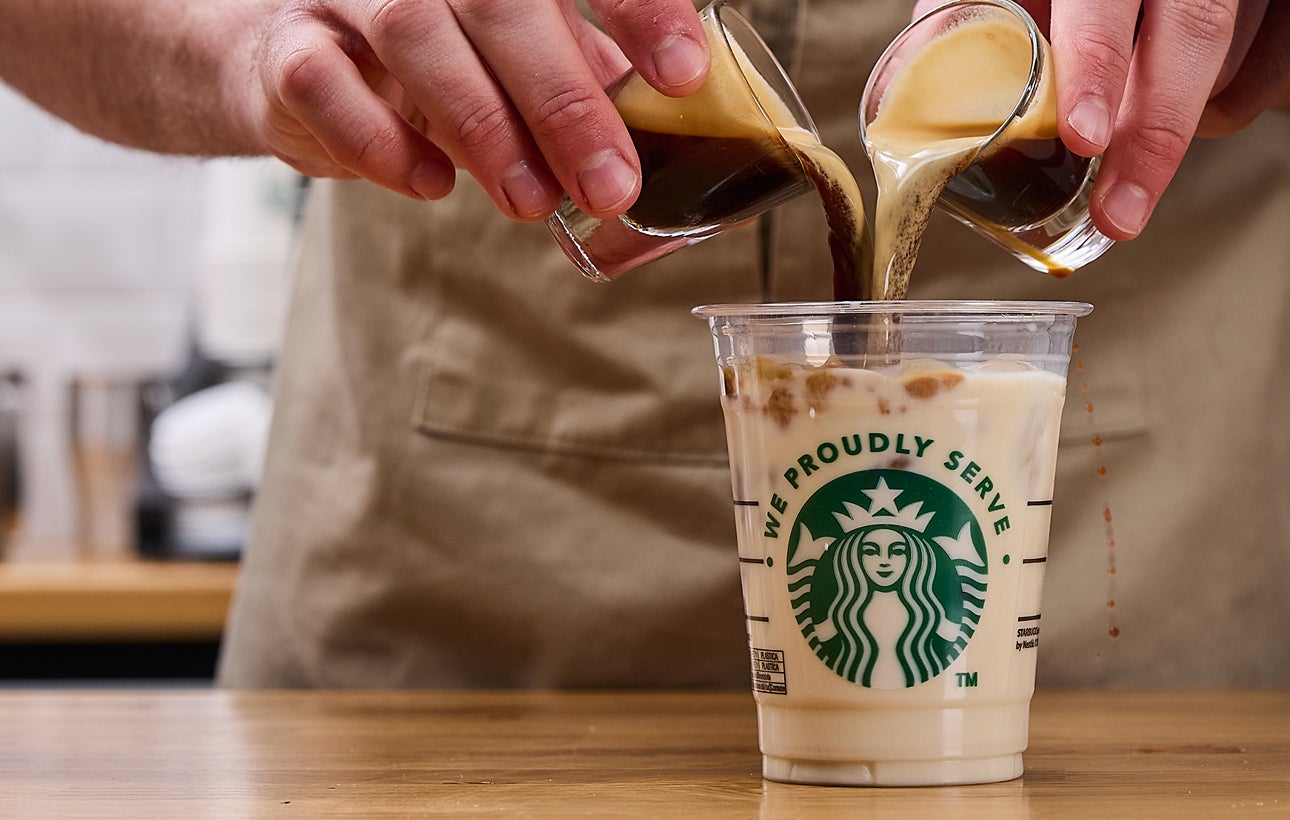 Un barista toarnă două shoturi de espresso simultan într-o cană We Proudly Serve Starbucks, umplută cu gheață și lapte.