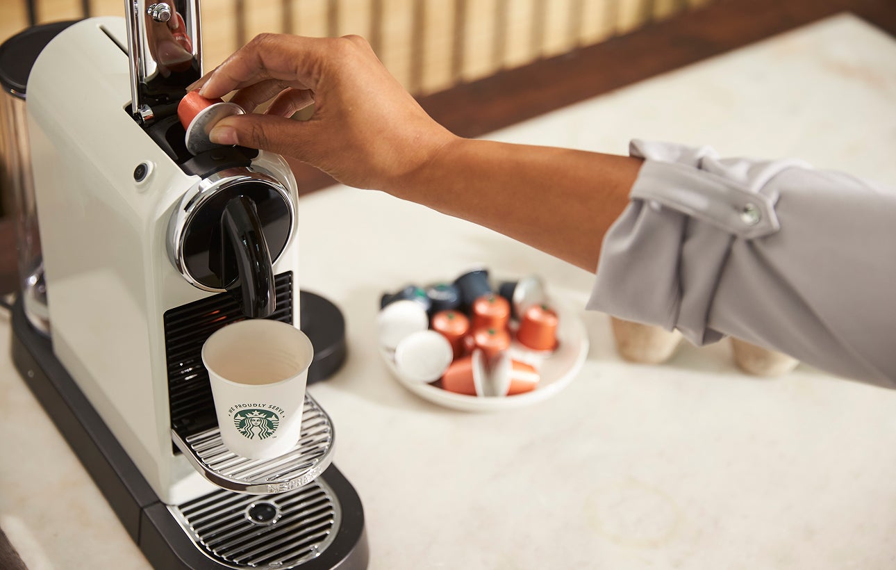 O persoană introduce o capsulă de cafea într-un aparat de cafea porționată, cu o cană We Proudly Serve Starbucks pe tava de s