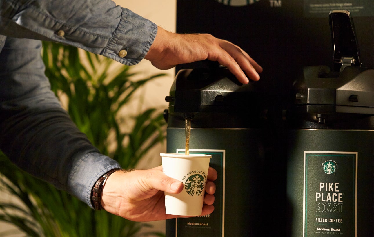 O persoană care servește cafea Starbucks Pike Place® Roast dintr-un distribuitor mare de cafea într-o ceașcă We Proudly Serve