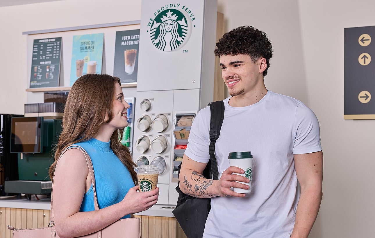 O femeie și un bărbat stând în fața unui aparat de cafea cu autoservire We Proudly Serve Starbucks
