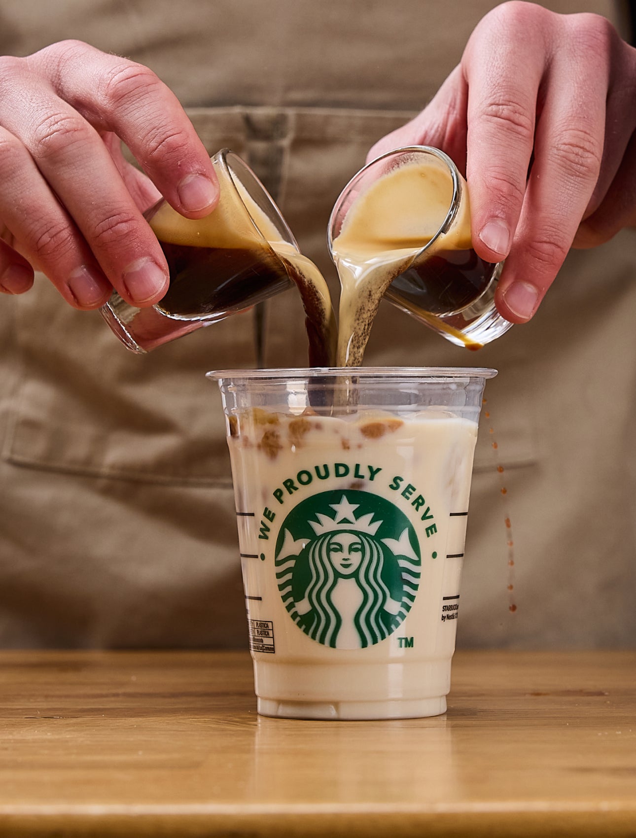 Un barista toarnă două shoturi de espresso simultan într-o cană We Proudly Serve Starbucks, umplută cu gheață și lapte.