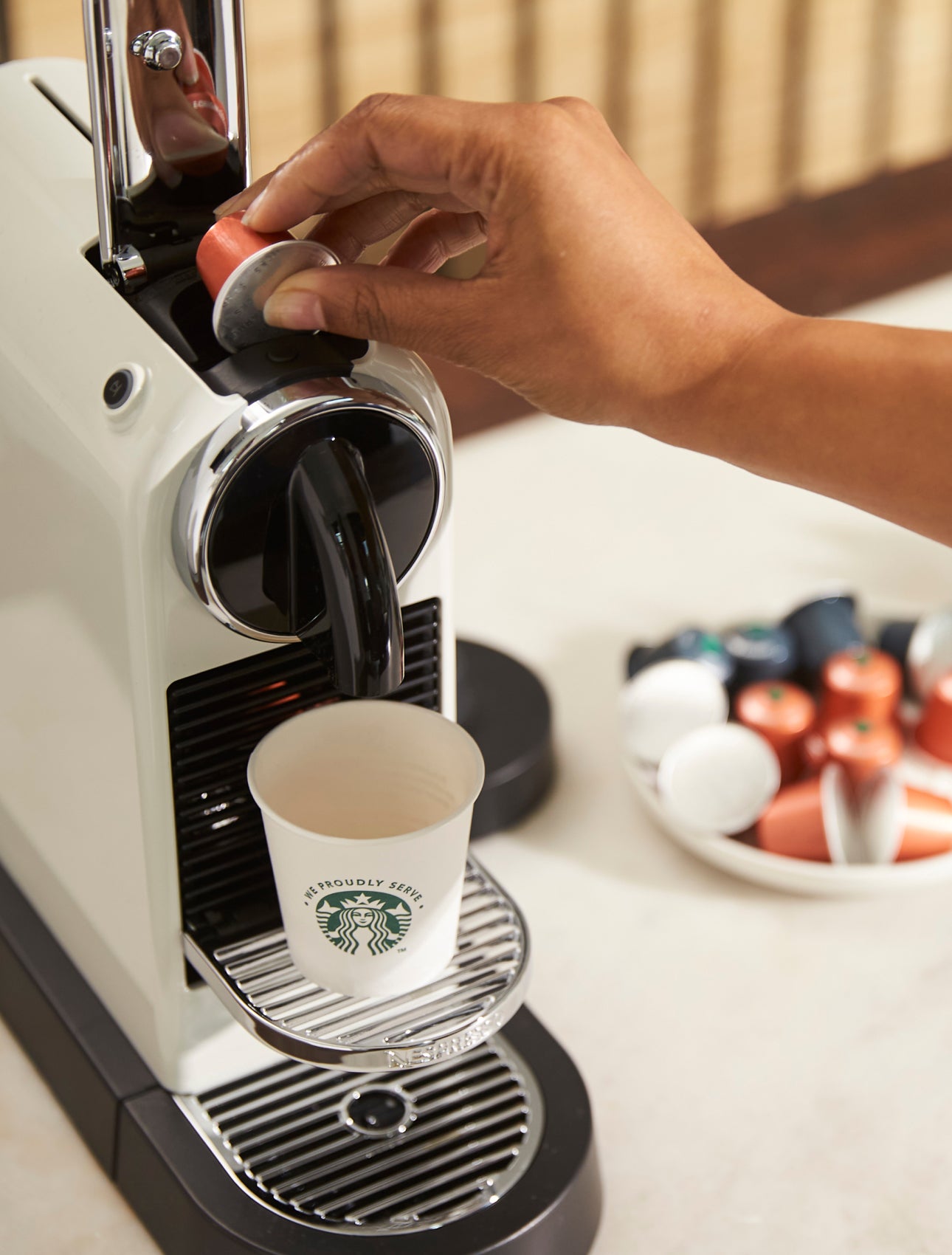O persoană introduce o capsulă de cafea într-un aparat de cafea porționată, cu o cană We Proudly Serve Starbucks pe tava de s