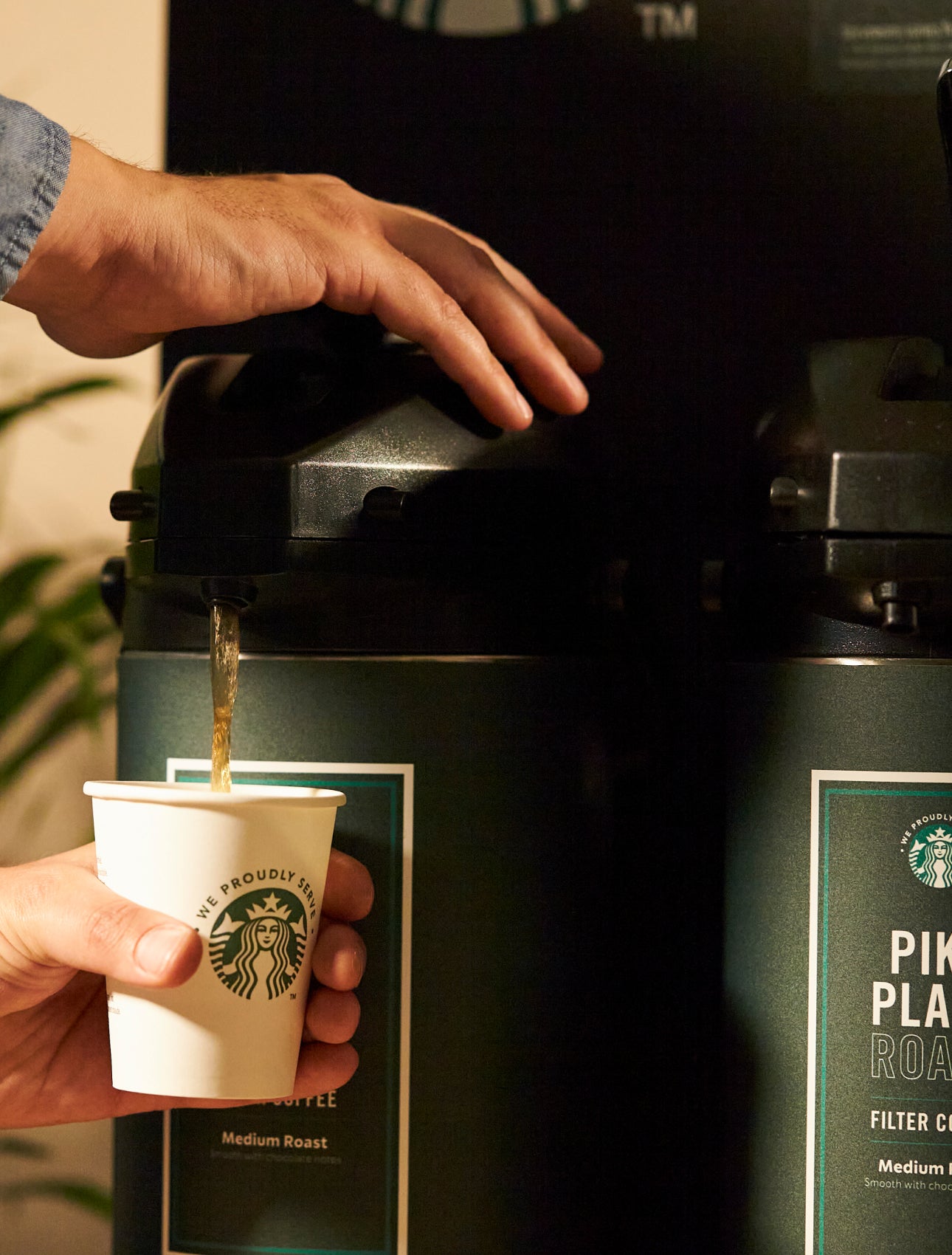 O persoană care servește cafea Starbucks Pike Place® Roast dintr-un distribuitor mare de cafea într-o ceașcă We Proudly Serve