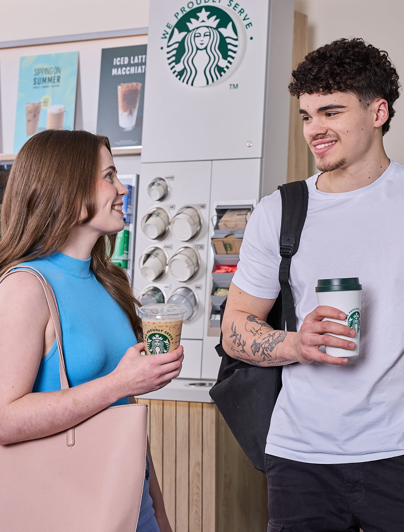O femeie și un bărbat stând în fața unui aparat de cafea cu autoservire We Proudly Serve Starbucks