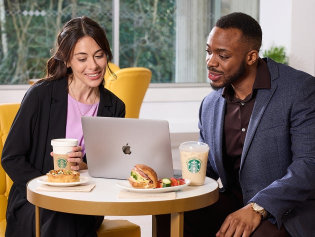 Două persoane așezate în fața unui laptop, cu cafea We Proudly Serve Starbucks și sandvișuri pe masă