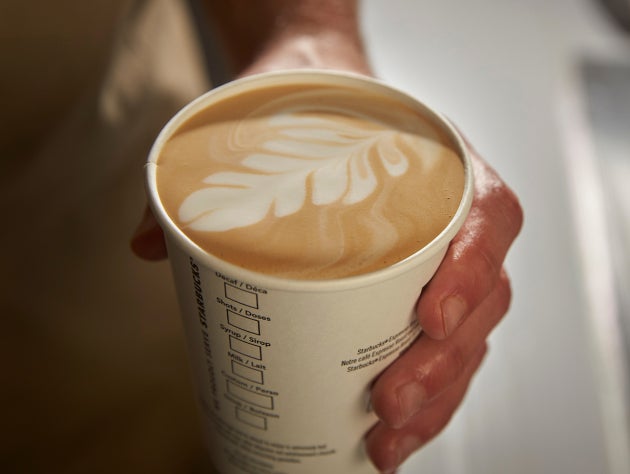  O mână care ține o ceașcă de cafea We Proudly Serve Starbucks, cu artă latte deasupra