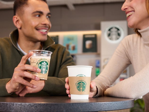 Două persoane care țin cu mândrie băuturi We Proudly Serve Starbucks într-o cafenea