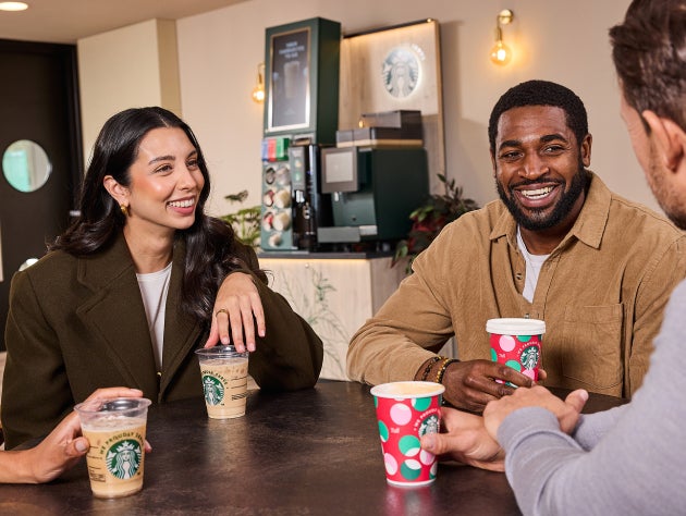 Patru persoane la o masă într-o cafenea, zâmbind și ținând în mână băuturi Starbucks în pahare festive și standard We Proudly