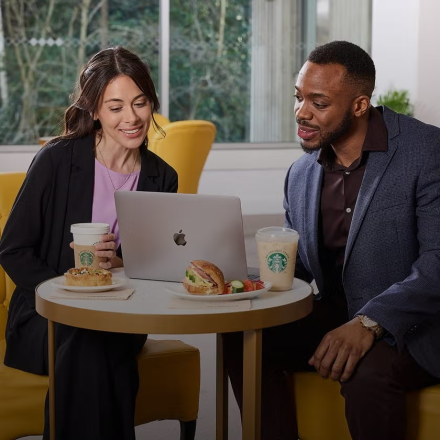 Două persoane așezate în fața unui laptop, cu cafea We Proudly Serve Starbucks și sandvișuri pe masă