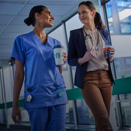 Doi profesioniști din domeniul sănătății mergând împreună într-o unitate medicală, zâmbind și ținând în mână băuturi We Proud