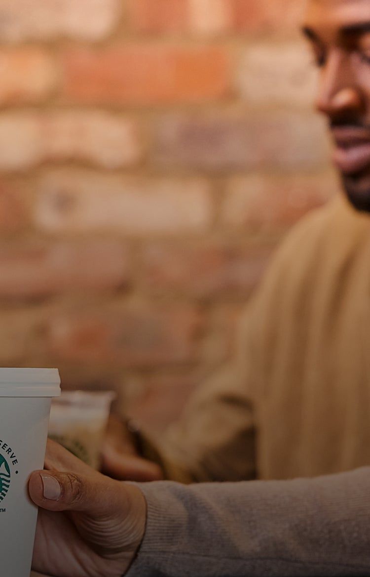 O persoană care ține o ceașcă We Proudly Serve Starbucks în fața unui fundal neclar de cărămidă