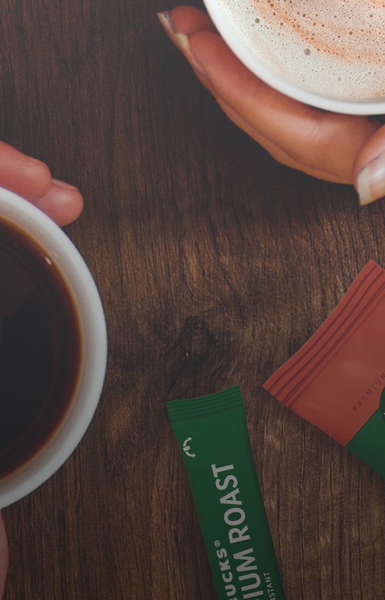 Două mâini țin o cafea neagră și un cappuccino spumos pe o masă, lângă pliculețe de cafea Starbucks® Premium Instant