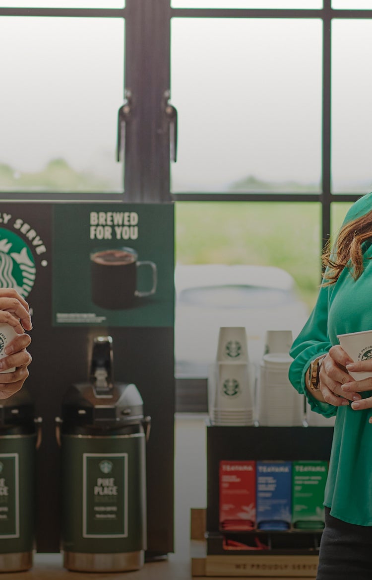 Două persoane care stau de vorbă, ținând în mână căni We Proudly Serve Starbucks, în fața distribuitoarelor de cafea la filtr