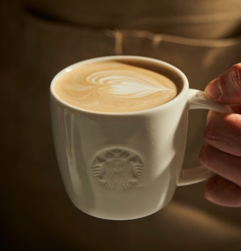 O mână care ține o cană albă Starbucks umplută cu un latte, acoperită cu artă latte în formă de inimă