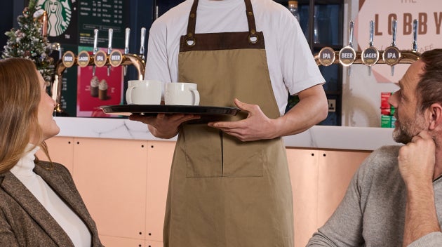 Un barista servește două băuturi Starbucks clienților la barul din holul hotelului, cu un meniu We Proudly Serve Starbucks® î