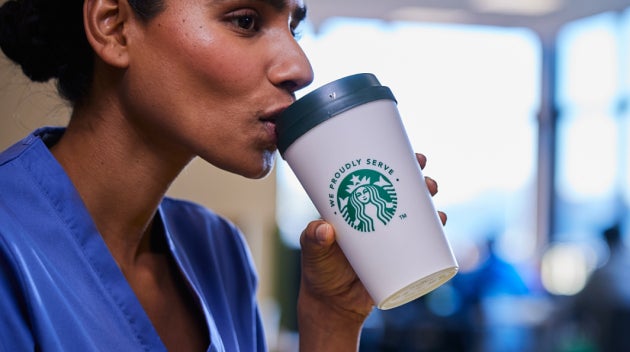 Un angajat din domeniul sănătății, îmbrăcat în halat albastru, bea o cafea We Proudly Serve Starbucks în timp ce stă în resta