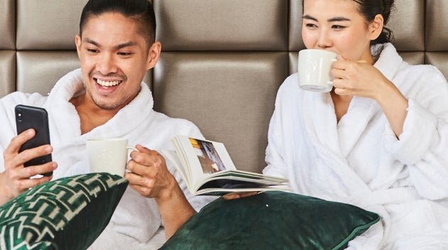 Un cuplu se relaxează pe un pat de hotel în halate albe, savurând cafea Starbucks. 