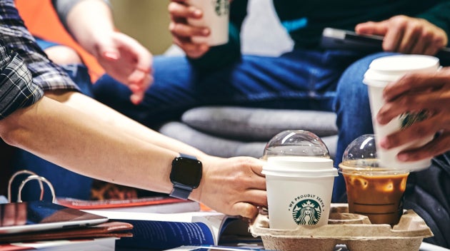 Un grup de studenți în jurul unei mese cu cărți și caiete, unul dintre ei luând o cafea We Proudly Serve Starbucks de pe masă