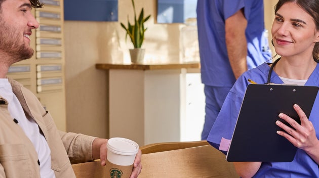 O femeie îmbrăcată în halat albastru vorbește cu un pacient ținând în mână o ceașcă de cafea We Proudly Serve Starbucks într-