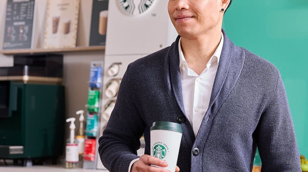 Angajat zâmbitor, cu cafea We Proudly Serve Starbucks și un laptop în mâini, în fața unui aparat de cafea cu autoservire